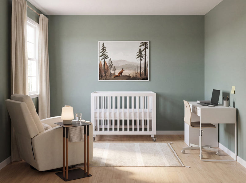 Small nursery office combo room in daylight: a white crib, a glider with a slim side table, a wall-facing desk, and a fox forest print above the crib
