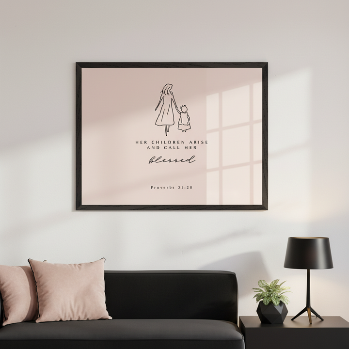 Modern living room with a black sofa and two pink pillows. A large framed wall art hangs above the sofa, featuring a minimalist line drawing of a mother and child, with the text "HER CHILDREN ARISE AND CALL HER blessed. Proverbs 31:28" on a pale pink background.