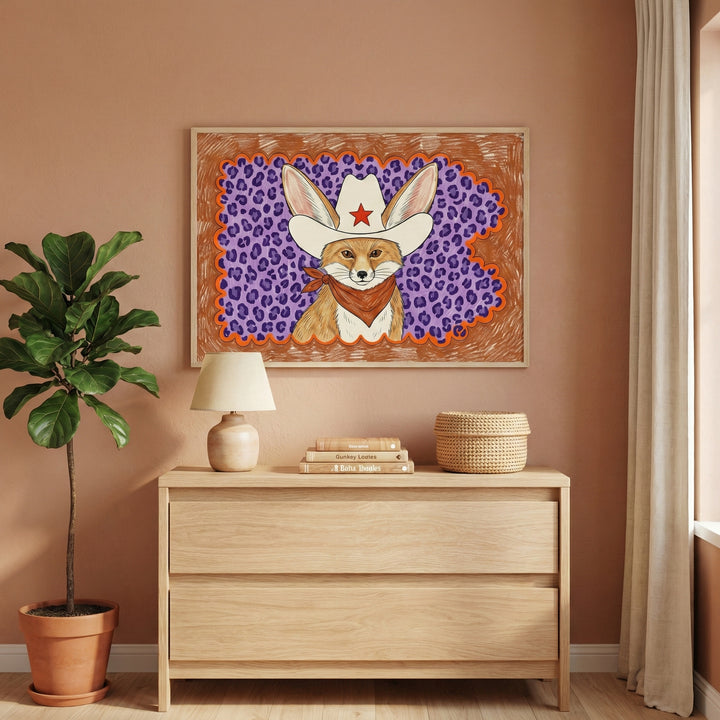 Cowboy fox wall art displayed above a wooden dresser in a warm-toned room with books and natural decor. The framed illustration's bold patterns and characterful fox design create an eye-catching focal point for playful or southwestern-inspired interiors.