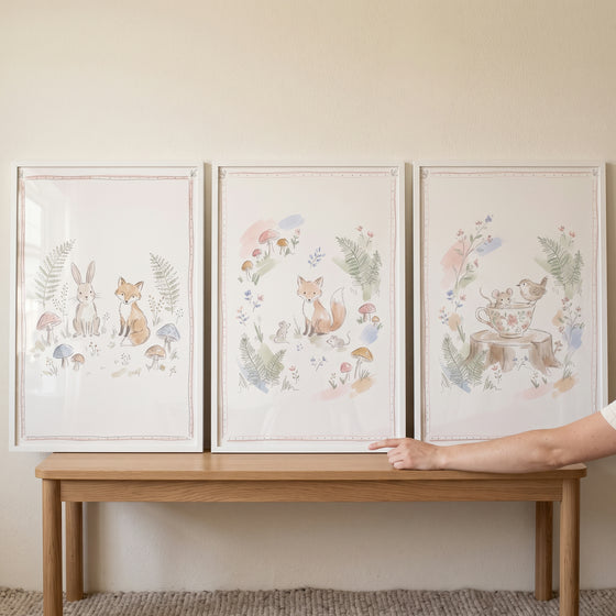 An airy charming woodland animals wall art set of 3 watercolor prints of foxes and rabbits on a light wooden bench.