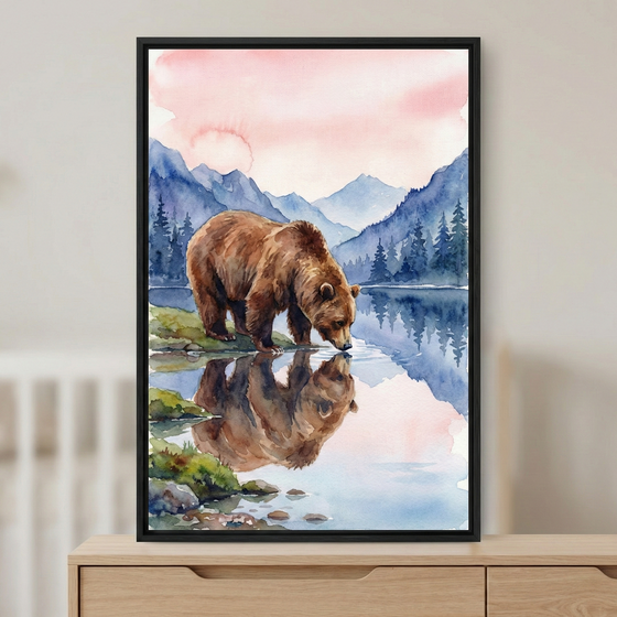 A serene watercolor artwork depicting a large brown bear at a lake's edge. The bear is captured in a profile view, leaning down to drink from the clear water, which perfectly mirrors its form. The landscape features rolling blue mountain peaks and a dense forest of evergreen trees under a soft, pastel pink and orange sky. This piece is presented in a slim black frame, resting on a light wood dresser. The style is defined by its fluid brushwork and gentle color palette.