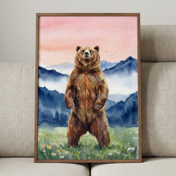A vertical watercolor artwork featuring a large brown bear standing tall on its hind legs in a grassy meadow dotted with small yellow and white flowers. Behind the bear, layers of mist-covered mountains transition from dark blue to soft indigo. The sky above is a delicate blend of pink and peach colors, with a few small bird silhouettes flying in the distance. The piece is enclosed in a simple brown wood frame and is shown leaning against a light grey textured sofa.