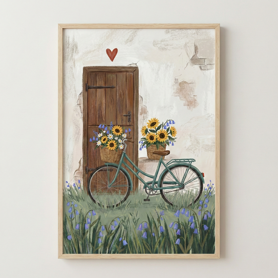 A framed piece of art featuring a rustic scene with a vintage-style teal bicycle parked in front of a dark brown wooden door. A small red heart is positioned above the door on a textured, off-white wall. Wicker baskets overflowing with sunflowers and blue and white blossoms are on the bicycle’s handlebars and on the ground nearby. The scene is set in a field of tall green grass and blue bell-shaped flowers. The artwork is shown in a light-toned natural wood frame.