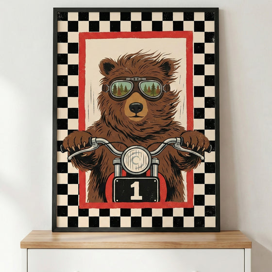 A framed illustration of a brown bear riding a motorcycle. The bear is wearing goggles that reflect a forest scene. Its paws are on the handlebars, and the motorcycle features a front headlight and a black number plate with the number one. The central image is surrounded by a red border and a wide black and white checkered pattern. The poster is in a black frame, sitting on a light-colored wooden surface.