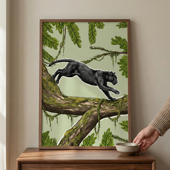 An illustration in a dark wood frame depicts a sleek black panther mid-leap across a moss-covered tree branch. Set against a pale green background, the scene is filled with lush tropical foliage, including various green leaves and hanging vines. The panther is rendered with a thin white outline that makes its form pop from the botanical surroundings. The framed piece sits on a wooden sideboard, and a hand is visible to the right, placing a small ceramic bowl nearby.
