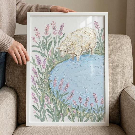 Framed painting of a textured white sheep standing at the edge of a soft blue pond surrounded by tall lavender stems rests against a cushioned chair while hands hold the frame. The pastel colors and thick brushstrokes create a calm gentle countryside scene.