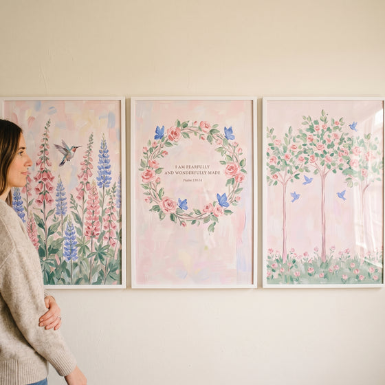 Woman views a blush pink meadow garden wall art set of 3 depicting flowers, hummingbirds, butterflies, and trees.