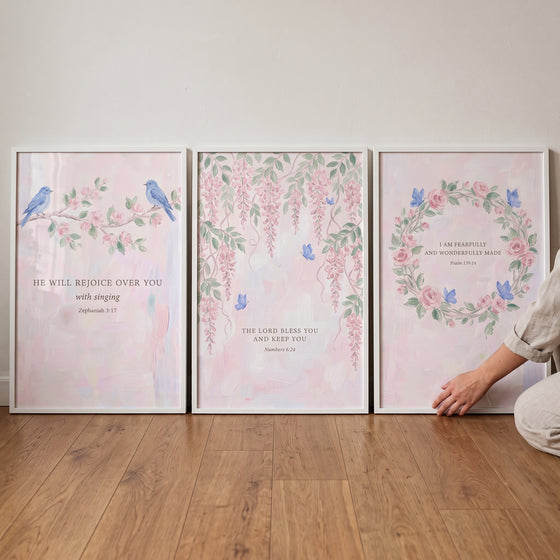 Botanical bible verse wall art set of 3 with floral designs and birds displayed in white frames on a wood floor.