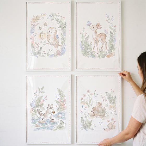 A person hangs a botanical wildflower woodland animals wall art set of 4 featuring an owl, deer, raccoon, and turtle.