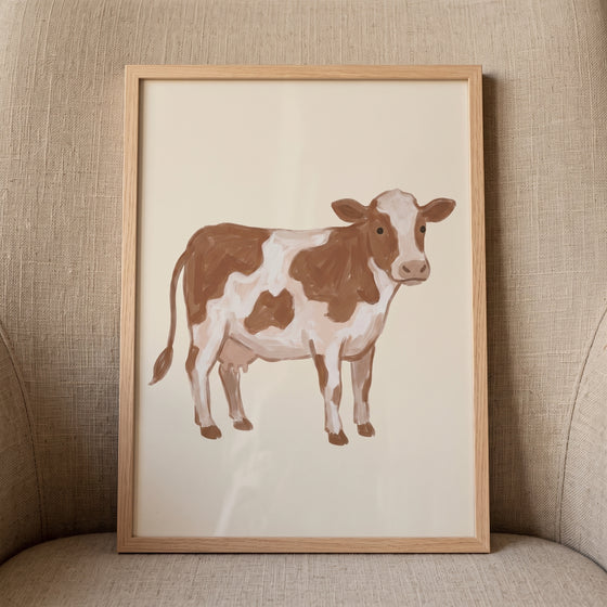 Brown white spotted cow wall art in a light wood frame rests on a textured beige armchair.