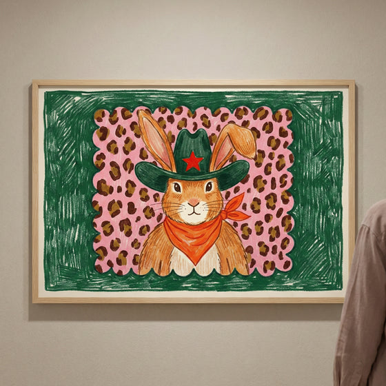 Framed wall art of a confident bunny in a dark green cowboy hat with a red star and orange bandana, posed in front of a pink and brown leopard print with a green crayon-style border. The quirky western bunny illustration adds a fun twist to animal-themed wall art.