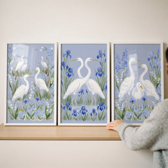 Coastal garden white egret bird wall art set of 3 shows white birds among blue flowers in frames on a wooden shelf.