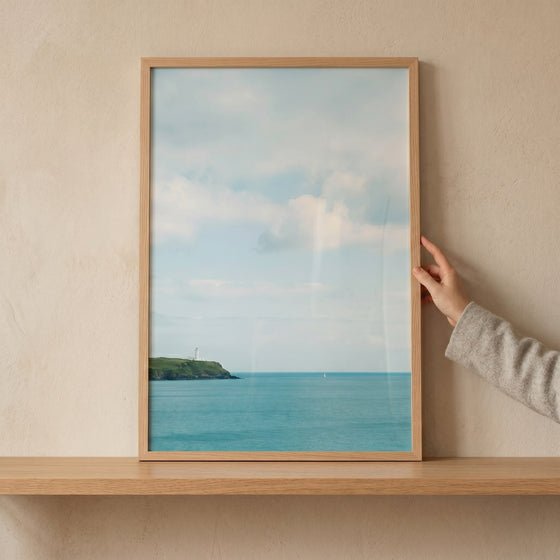 Hand holds wood framed coastal turquoise sea lighthouse wall art on a shelf against a neutral wall.