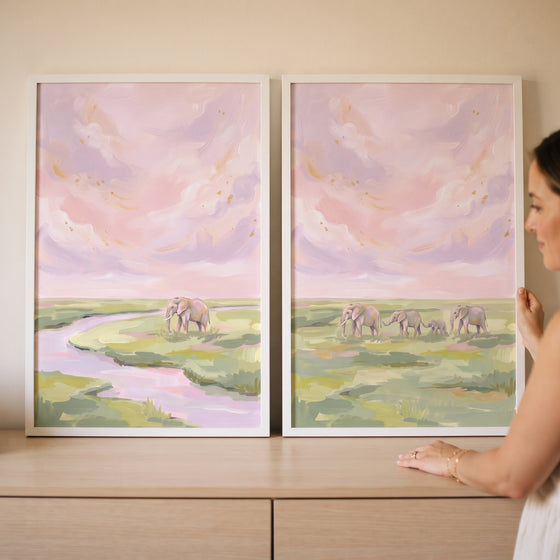 A woman holds a coquette blush pink elephant wall art set of 2 featuring elephants by a river in a pastel savanna.