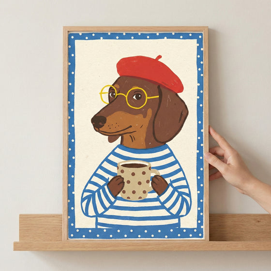 A whimsical illustration of a dachshund wearing a red beret, round yellow glasses, and a blue and white striped shirt. The dog holds a beige polka-dot mug between its paws. The artwork is set within a blue border with white polka dots and is displayed in a light wood frame on a matching shelf. A hand is visible touching the side of the frame.