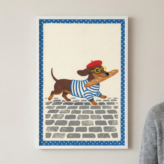 An illustration of a brown dachshund trotting across a cobblestone street. The dog is wearing a red beret, round yellow glasses, and a blue and white striped shirt, and it carries a long loaf of bread in its mouth. The artwork is set against a cream background with a blue and white polka-dot border, all contained within a simple white frame.