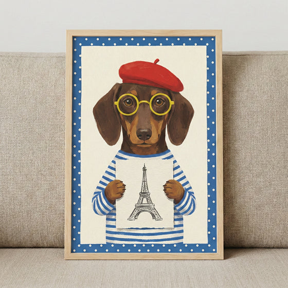 A framed illustration featuring a whimsical portrait of a dachshund. The dog is dressed in a red beret, circular yellow-framed glasses, and a blue and white striped top. It holds a small piece of paper with a drawing of an iconic Parisian tower on it. The image is surrounded by a blue border with white polka dots, encased in a light wood frame, and positioned against a neutral-toned fabric sofa.