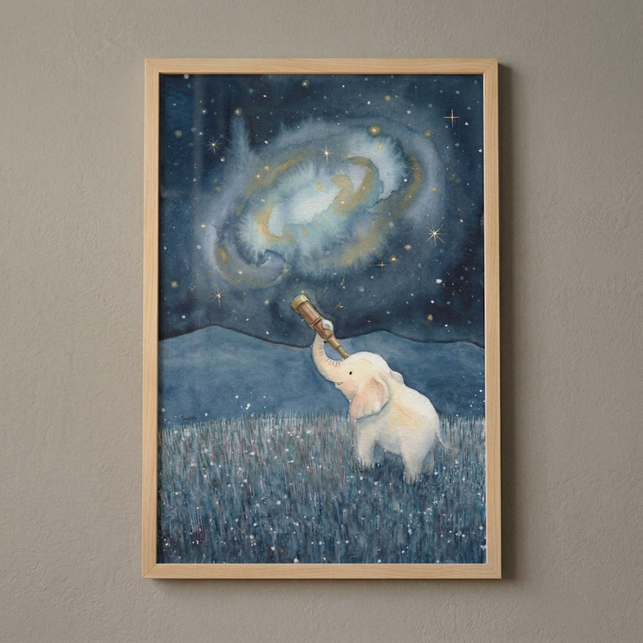 A framed watercolor-style illustration shows a small, white cartoon elephant in a field of dark blue grass with tiny white dots. The elephant holds a telescope with its trunk, looking up at a vast night sky. Above it, a large, glowing galaxy swirls in soft blue and gold tones, surrounded by many twinkling stars. Silhouetted mountains are visible in the distance. The artwork is held in a light wooden frame against a neutral-colored wall.