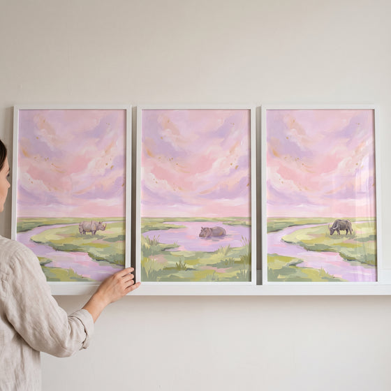 Hand adjusts a dusk sky safari animals wall art set of 3 with a rhino, hippo, and buffalo in pastel landscapes.