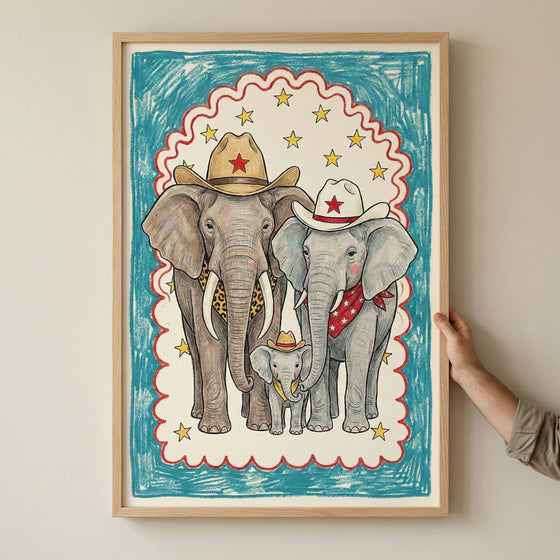 Framed illustration of three elephants dressed in western cowboy attire, including hats with red stars, a leopard print scarf, and a red bandana, with a colorful border of stars and blue scribble background. A hand is holding the frame against a neutral wall.