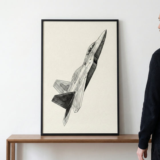 A framed black and white pencil sketch of a modern fighter jet soaring diagonally upwards. The detailed illustration features intricate shading and is presented on off-white paper within a thin black frame. The artwork sits on a wooden table against a plain white wall, with a person partially visible on the right.