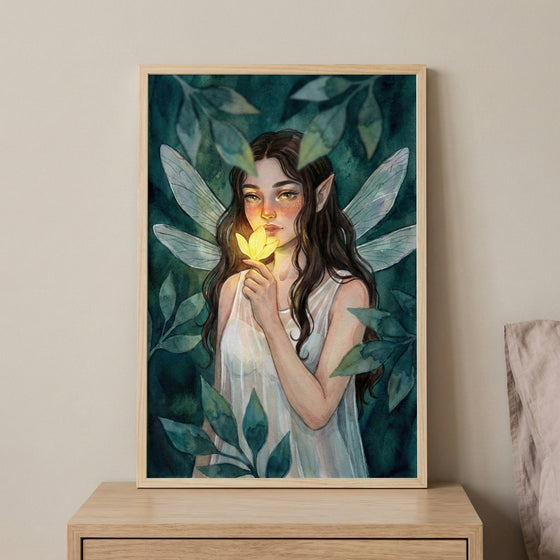 A framed artwork featuring a young woman with pointed ears and delicate, translucent wings. She has long dark hair and is holding a glowing yellow flower near her face. She is wearing a white sleeveless dress. The background is filled with various shades of green and teal leaves. The frame is made of light wood and is resting on a wooden surface against a plain wall.