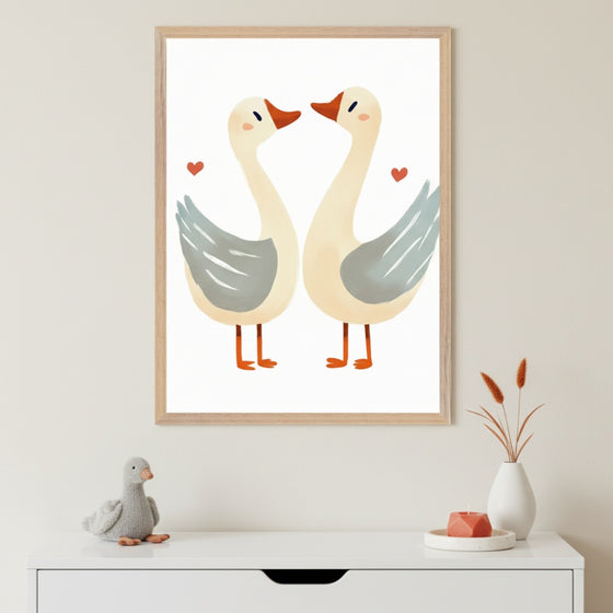 Neutral-toned room with a white dresser. Above it hangs a framed illustration of two stylized geese, facing each other, with red hearts. A plush gray goose, a white vase with dried plants, and a coral candle are on the dresser.
