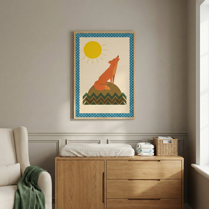 A serene nursery setting with a framed art print centered on a neutral-colored wall. The print features a graphic illustration of an orange fox sitting on a green hill and howling at a large yellow sun, set against a cream background with a blue and white polka-dot border. Below the art, a light wood dresser holds a changing mat, diapers, and a small wicker basket. A cream-colored armchair with a draped green blanket is partially visible on the left, and soft light enters from a window on the right.