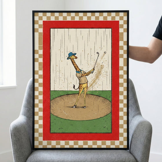 A framed illustration of a giraffe dressed in a blue hat and beige shirt playing golf. The giraffe is mid-swing in a sand trap, with sand and a golf ball flying into the air while rain falls in the background. The central artwork is enclosed by a bold red border and an outer tan and white checkered pattern. The piece is in a black frame and sits on a grey armchair.