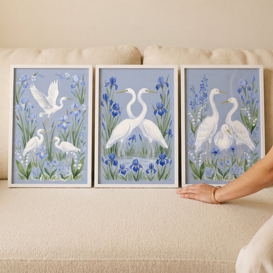 Grandmillennial blue iris bird egret wall art set of 3 depicting white egrets and flowers, displayed on a cream sofa.