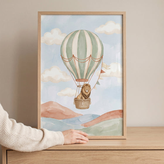 Hot air balloon lion wall art in a wood frame shows a lion in a striped balloon over watercolor hills and clouds.