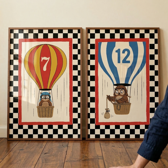 Two vertical art prints in simple wooden frames lean against a plain, light-colored wall on a polished wooden floor. Both prints feature a whimsical illustration framed by a wide black-and-white checkered outer border and a thinner red inner border.
The print on the left shows a hot air balloon with alternating red and yellow vertical stripes, with a large white number "7" displayed prominently.