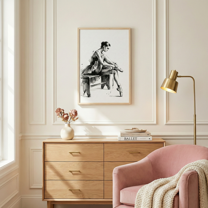 A minimalist and elegant interior scene featuring a framed piece of artwork on a cream-colored wall with classic decorative molding. The vertical print, set in a light natural wood frame, shows a black and white ink sketch of a ballerina in a tutu, sitting on a bench and adjusting her shoe.