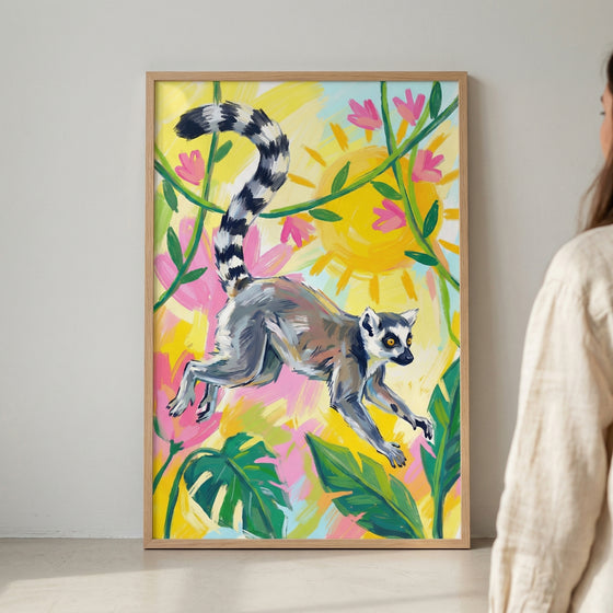 A framed piece of vibrant artwork displays a ring-tailed lemur in a mid-air leap. The lemur, with its characteristic long, black-and-white banded tail, is the central focus against a bright, colorful background. Stylized tropical elements like large green leaves, pink flowers, and a glowing yellow sun surround the animal. The style features bold, expressive brushstrokes and a playful use of color. The art is presented in a simple wooden frame, leaning against a light-colored wall.