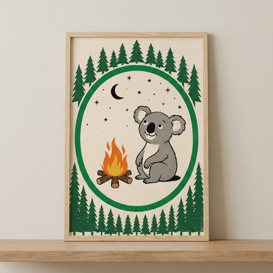 A framed illustration of a smiling cartoon koala sitting next to a small campfire. The central image is set against a cream background and enclosed within a thick green circular border. Above and below the circle are rows of dark green stylized evergreen trees. Inside the circle, a black crescent moon and several small stars are scattered in the sky. The artwork is held in a light wood frame resting on a natural wood surface against a neutral wall.