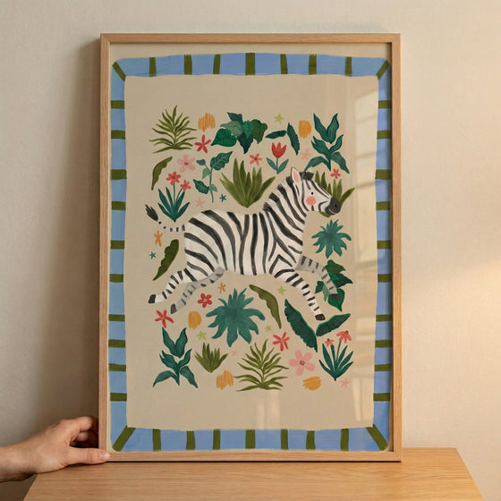 Framed zebra illustration with black and white stripes surrounded by colorful leaves and flowers, styled as playful safari nursery wall art. A hand steadies the frame on a wooden surface, showing scale and placement for decorating a child’s room.