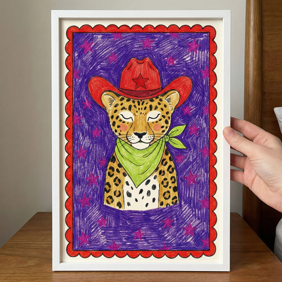 Colorful framed illustration of a leopard wearing a red cowboy hat with a star and a green bandana, set against a purple background with pink stars and a red scalloped border. A hand holds the framed art above a wooden surface.