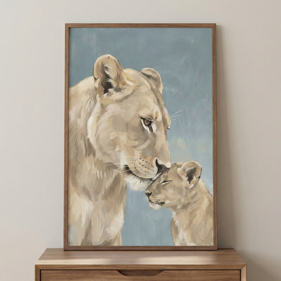 A vertical framed artwork captures a mother lion gently nuzzling her cub. The large lioness leans down, her nose softly touching the forehead of the cub, who has its eyes closed in a moment of peace. The illustration features visible, layered strokes in a palette of cream, sand, and light brown. The background is a flat, muted blue-gray. The piece is housed in a simple wooden frame and displayed on a light brown wooden tabletop against a plain, off-white wall.