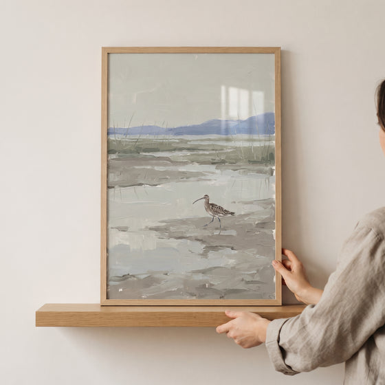 Hands place a framed long bill marsh curlew bird wall art on a wooden shelf featuring a muted coastal landscape.