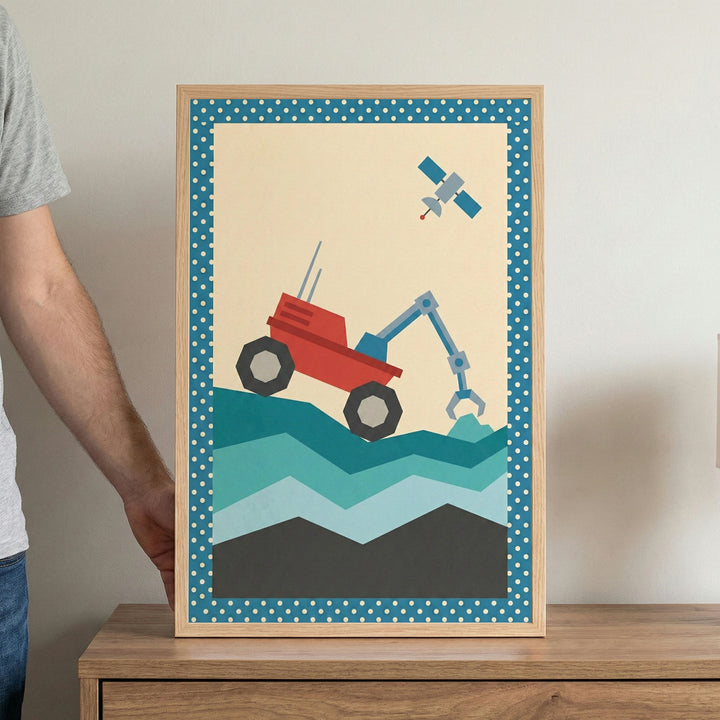 A minimalist, geometric illustration is displayed in a light wood frame, held by a person on a wooden surface. The artwork features a red space exploration rover with a blue mechanical arm perched on a jagged, multi-toned teal and grey landscape. Above the rover, a small blue and grey satellite floats in a beige sky. The style uses flat colors and simple shapes to create a modern, stylized scene.