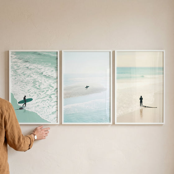 Minimalist pastel coastal surfer wall art set of 3 shows beach scenes as a hand reaches to adjust one of the frames.
