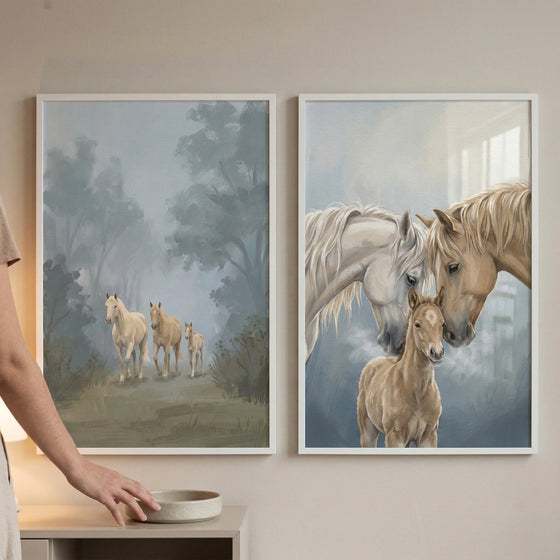 Two framed horse prints hang on a beige wall above a light surface, with part of a person visible at the left for scale. One print shows three pale horses walking through a misty wooded path, while the other shows two adult horses nuzzling a foal against a soft blue gray background.