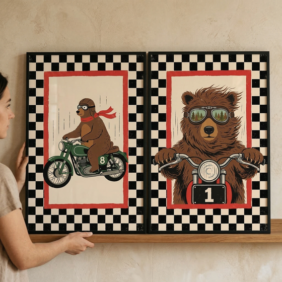 A person is seen from the side, holding two rectangular framed art prints onto a wooden shelf. The prints feature vintage-style illustrations of brown bears as motorcyclists.
The print on the left shows a profile view of a brown bear riding a green motorcycle. The bear is wearing a brown leather cap, goggles, and a long red scarf that flows behind it, suggesting movement. Small vertical lines in the background further enhance the sense of speed.