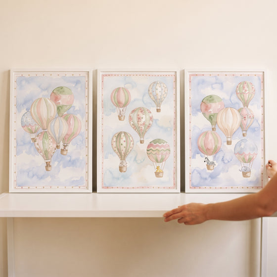 Muted hot air balloon wall art set of 3 features soft pastel balloons and animals in white frames on a white shelf.
