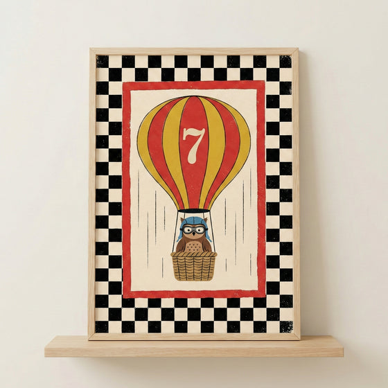 A framed wall art print features a whimsical illustration of a red and yellow striped hot air balloon with a large white number seven on its side. In the woven basket below, a small brown owl wears a blue aviator cap and goggles. The central image is surrounded by a solid red border, which is enclosed by a bold black and white checkerboard pattern. The artwork is presented in a light-colored wood frame and sits on a matching wooden shelf against a neutral wall.