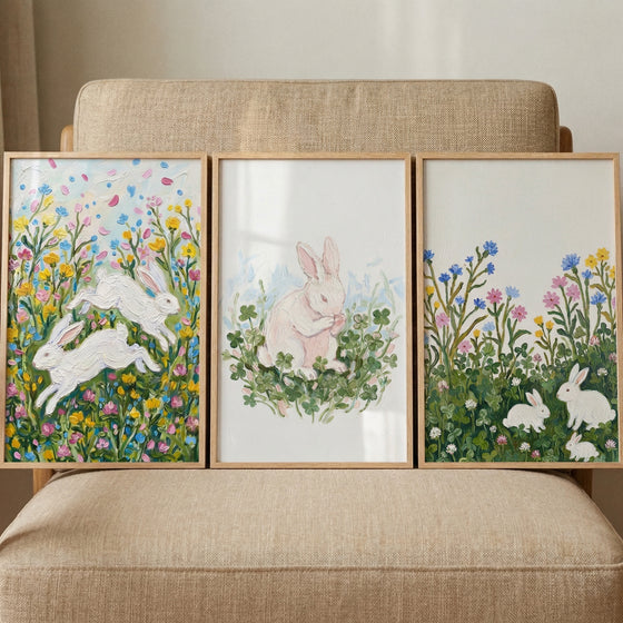 A set of three framed, textured paintings resting on a beige armchair. The artwork features white bunnies in different nature scenes: leaping through colorful wildflowers on the left, sitting among green clovers in the center, and resting in a floral meadow on the right.