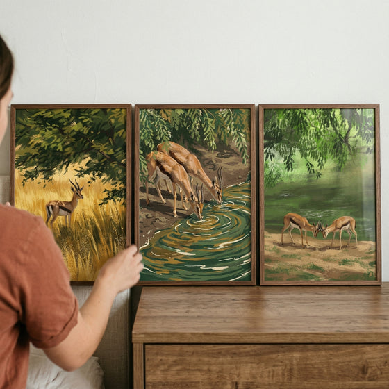 A hand touches a painterly green savanna gazelle wall art set of 3 featuring wild animals by trees and a water source.