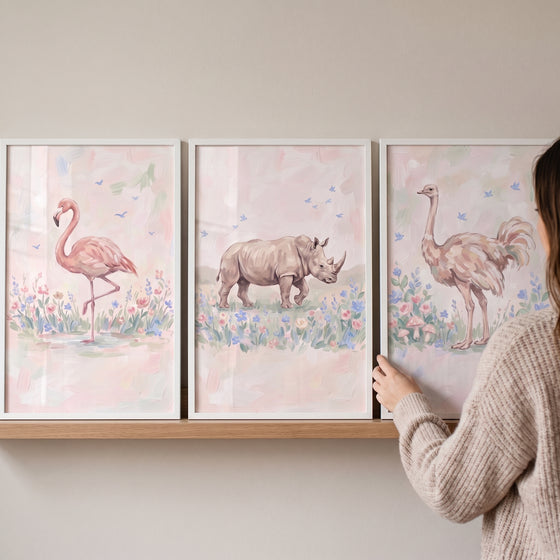 A woman hangs a pastel meadow safari animals wall art set of 3 featuring a flamingo, rhino, and ostrich in white frames.