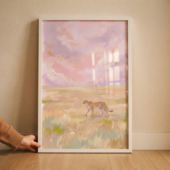 A hand holds a framed pastel pink sky cheetah animal wall art piece of a cheetah walking through a golden field.