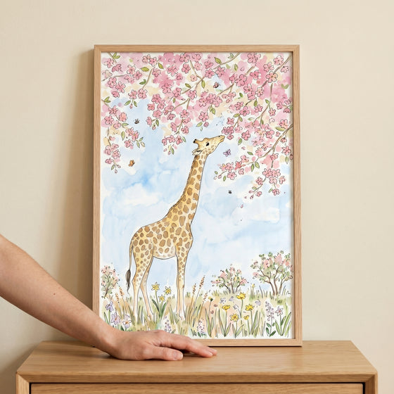 A hand touches a wooden frame containing pink blossom giraffe wall art of a giraffe under a blooming cherry tree.